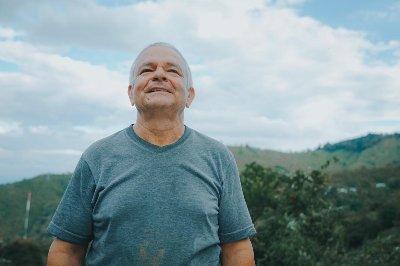 Aparece un señor, adulto mayor, de medio cuerpo, al aire libre, sonriente, con una camiseta color azul y de fondo las montañas verdes.