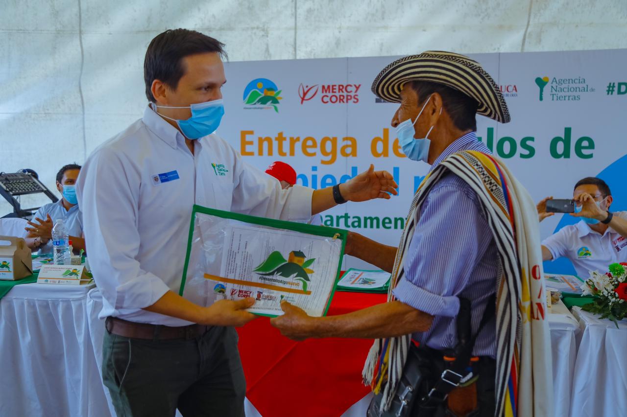 Funcionario de la ANT, vestido de camisa blanca, pantalón azul y tapabocas, hace entrega título de propiedad a un campesino, quien lleva un sombrero, poncho, carriel y tapabocas. De fondo en mesa cubierta con mantel blanco otros funcionarios de la ANT y una pancarta de la agencia.