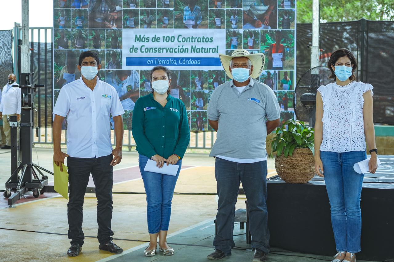 La Directora de la ANT, Myriam Martínez Cárdenas, aparece de pie y cubriendo sus bocas con tapabocas, con tres personas más, campesinos. De fondo una pancarta con varias fotografías de campesinos beneficiados de la entrega de títulos.