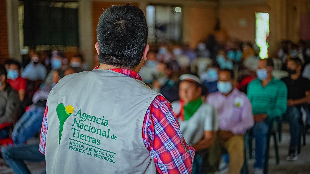 Fotografía de Audiencia Pública para la creación de la Zona de Reserva Campesina – ZRC de Santa Rosa, en Cauca. En ella aparece dando la espalda un funcionario de la ANT con su chaleco corporativo, hablando frente a varios campesinos quienes están sentados en sillas de plástico color azul.