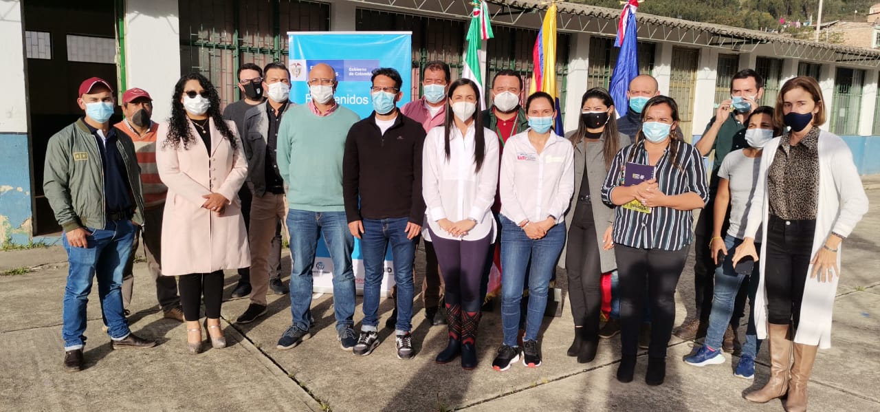 Diez hombres y siete mujeres posan de pie para la toma de una fotografía. De fondo una vaya vertical con el logo del Gobierno, y tres banderas entre ellas la de Colombia. 