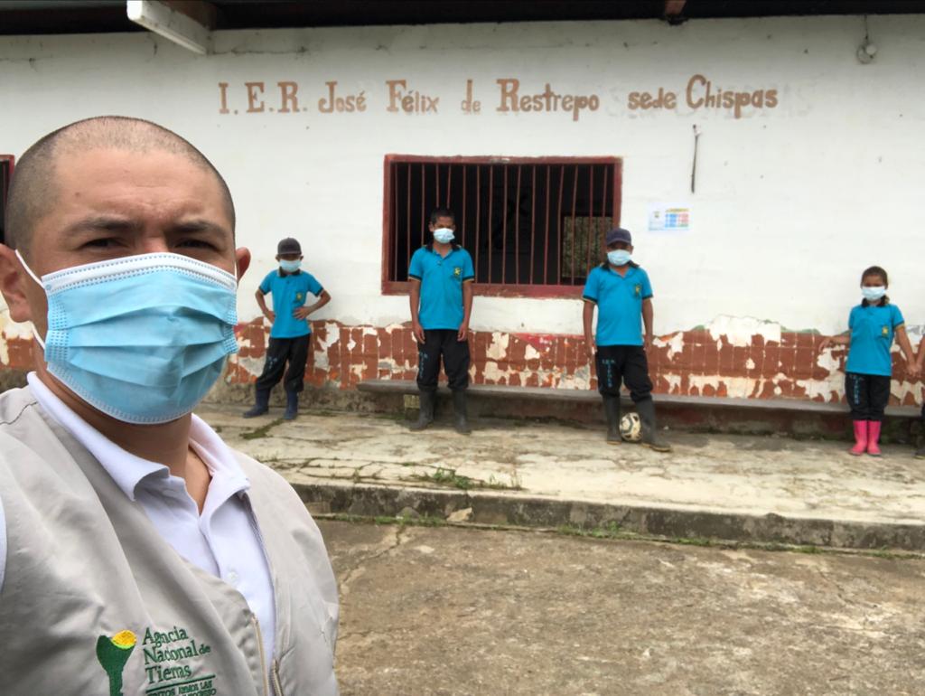 Selfi tomada por un hombre funcionario de la ANT. Él de medio cuerpo, luce el chaleco corporativo de la ANT y lleva puesto camisa blanca y tapabocas. De fondo aparece la institución educativa José Félix Restrepo sede Chispas y cuatro niños estudiantes vistiendo todos camisas iguales de color azul, pantalón negro y botas de caucho. 