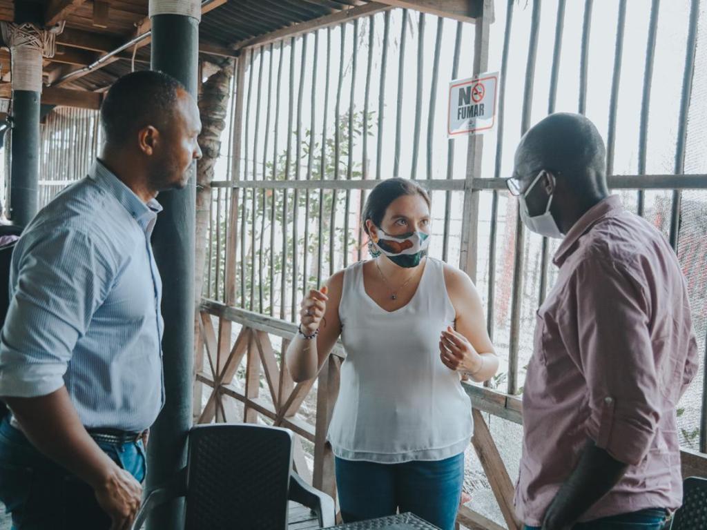 La Directora de la ANT, Myriam Martínez Cárdenas, aparece de pie dialogando con dos hombres afrodescendientes en una casa hecha de madera.