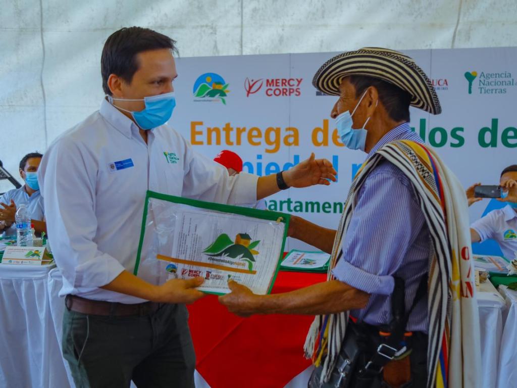 Funcionario de la ANT, vestido de camisa blanca, pantalón azul y tapabocas, hace entrega título de propiedad a un campesino, quien lleva un sombrero, poncho, carriel y tapabocas. De fondo en mesa cubierta con mantel blanco otros funcionarios de la ANT y una pancarta de la agencia.