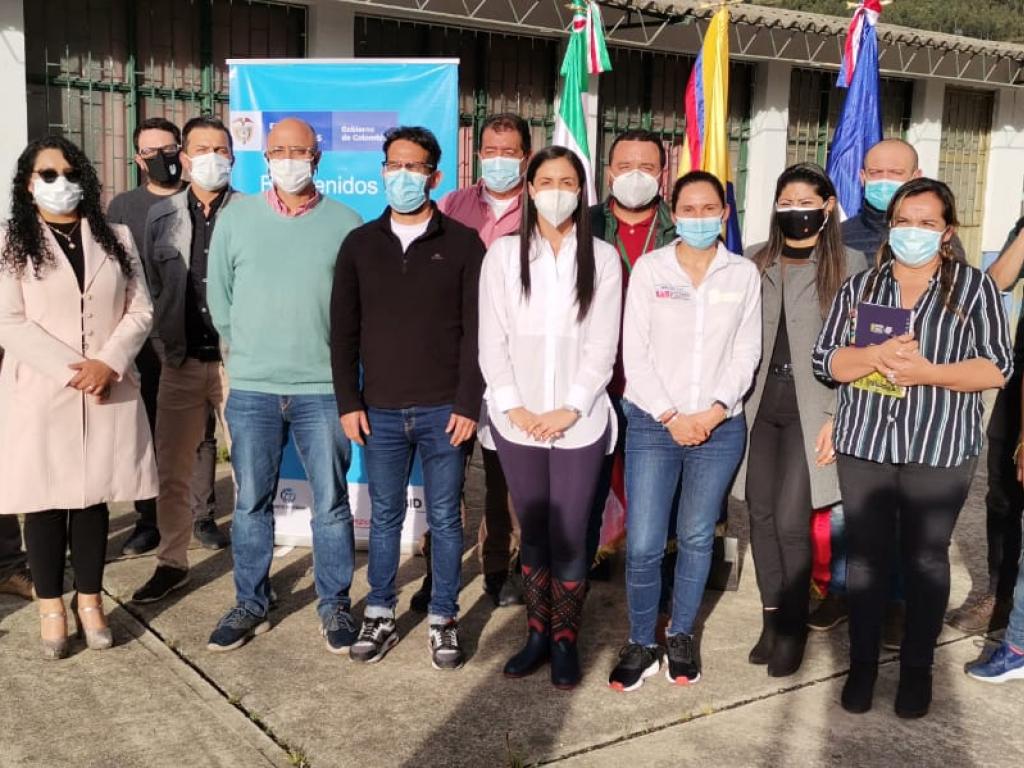 Diez hombres y siete mujeres posan de pie para la toma de una fotografía. De fondo una vaya vertical con el logo del Gobierno, y tres banderas entre ellas la de Colombia.